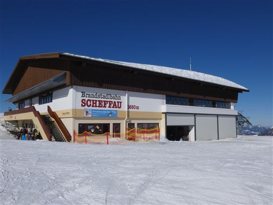 Ein großes Gebäude mit dem Namen „Brandstätterbahn Scheffau“ in einem verschneiten Berggebiet. Der klare blaue Himmel und der Schnee schaffen eine winterliche Atmosphäre.