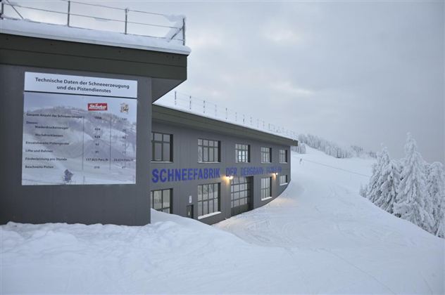 Ein modernes Gebäude in einer verschneiten Landschaft. Schilder mit technischen Informationen zur Schneeproduktion sind sichtbar.
