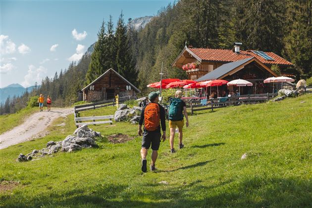 Zwei Wanderer gehen auf einem Weg zu einer Berghütte mit roten Sonnenschirmen. Die Umgebung ist grün und von Bäumen umgeben.