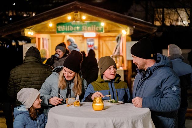 Eine Gruppe von vier Personen steht an einem runden Tisch und genießt Essen und Trinken. Im Hintergrund ist eine beleuchtete Verkaufsbude zu sehen.