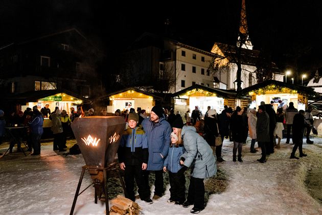 Ein festlicher Weihnachtsmarkt in den Bergen bei Nacht, umgeben von Schnee und beleuchtet von warmen Lichtern. Menschen versammeln sich um Feuerstellen, während im Hintergrund eine Kirche sichtbar ist.