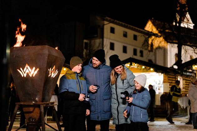 Eine Familie steht in einem festlich beleuchteten Markt und genießt warme Getränke. Im Hintergrund brennen Flammen und es sind festliche Stände zu sehen.