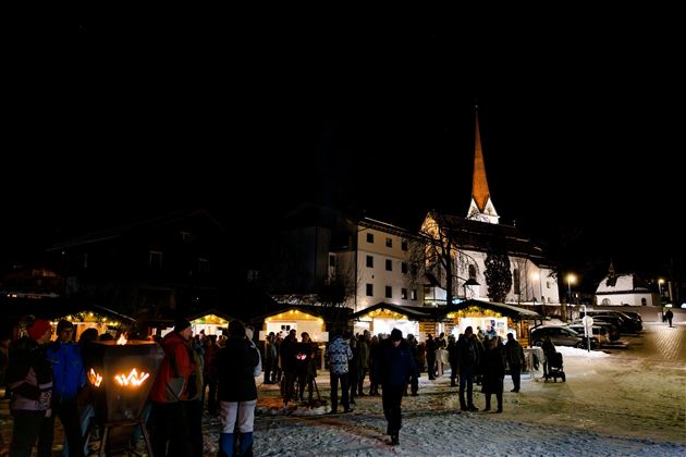 Ein festlicher Weihnachtsmarkt bei Nacht mit vielen Besuchern und stimmungsvollem Licht. Im Hintergrund ist eine Kirche mit einem hohen, spitzen Turm zu sehen.