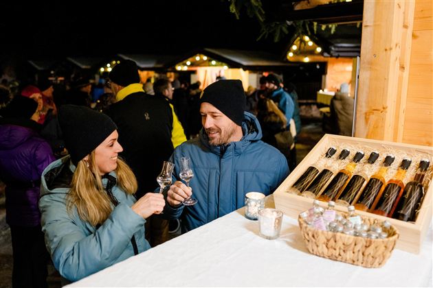 Ein Paar stößt mit Gläsern an und genießt die festliche Atmosphäre eines Marktes bei Nacht. Im Hintergrund sind weitere Menschen und stimmungsvolle Lichter zu sehen.