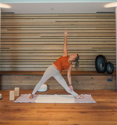 A woman is doing a yoga pose in a bright, modern room. The wooden walls and mats create a relaxing atmosphere.