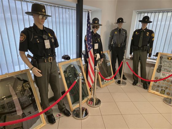 Die Ausstellung zeigt Figuren in Polizeiuniformen neben Rahmen mit Abzeichen und Erinnerungsstücken. Im Hintergrund ist eine Fahne zu sehen, die die Ermittlungsbehörde repräsentiert.