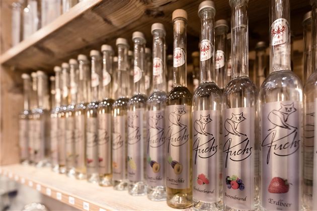 A series of clear bottles filled with various fruit brandies stands on a wooden shelf. The bottles are nicely labeled and showcase a variety of fruit aromas.
