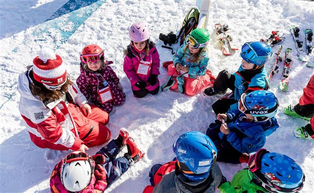 Eine Gruppe von Kindern sitzt im Schnee und hört einem Skilehrer zu. Rundherum sind Skiausrüstungen und verschneite Berge sichtbar.