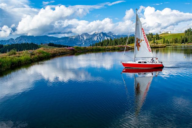 Ein rotes Segelboot schwimmt auf einem ruhigen See. Im Hintergrund sind Berge und eine schöne Wolkenlandschaft zu sehen.