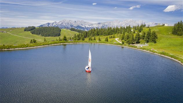 Ein malerischer See umgeben von grünen Wiesen und Bergen im Hintergrund. Ein Segelboot gleitet ruhig über das Wasser.