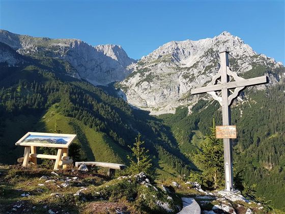Eine schöne Berglandschaft mit hohen Gipfeln und grünem Wald. Im Vordergrund steht ein Holzkreuz und eine Informationstafel.