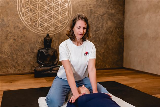 Eine Frau gibt eine Massage in einem ruhigen Raum. Im Hintergrund steht eine Buddha-Statue und ein Mandala ist an der Wand zu sehen.