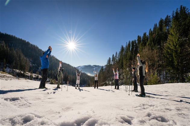 Eine Gruppe von Menschen macht eine Dehnübung im Schnee. Die Sonne scheint hell in den klaren blauen Himmel.
