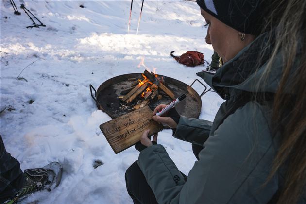 Eine Person sitzt im Schnee und schreibt auf einem Holzstück. Im Hintergrund brennt ein kleines Feuer.
