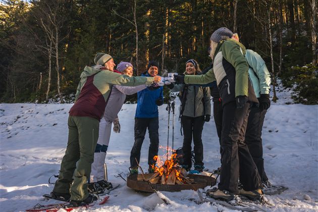 Eine Gruppe von sechs Menschen steht im Schnee um ein Lagerfeuer und stoßt mit Getränken an. Im Hintergrund sind Bäume und eine winterliche Landschaft sichtbar.
