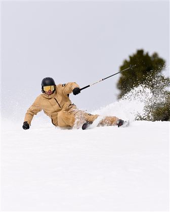 Ein Skifahrer fährt in einem verschneiten Gebiet. Er trägt eine beige Skijacke und einen Helm.