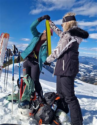 Zwei Personen bereiten sich im Schnee auf das Skifahren vor. Sie überprüfen ihre Ausrüstung vor einer malerischen Berglandschaft.