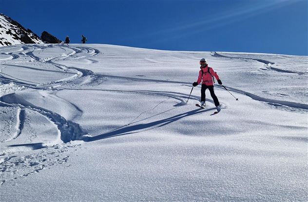 Eine Skifahrerin in pinker Kleidung fährt über eine schneebedeckte Piste. Im Hintergrund sind weitere Skifahrer und ein strahlend blauer Himmel zu sehen.