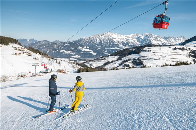 Zwei Skifahrer stehen auf einer verschneiten Piste und sehen auf die beeindruckenden Berge. Im Hintergrund ist eine Gondel zu sehen, die über die Landschaft schwebt.