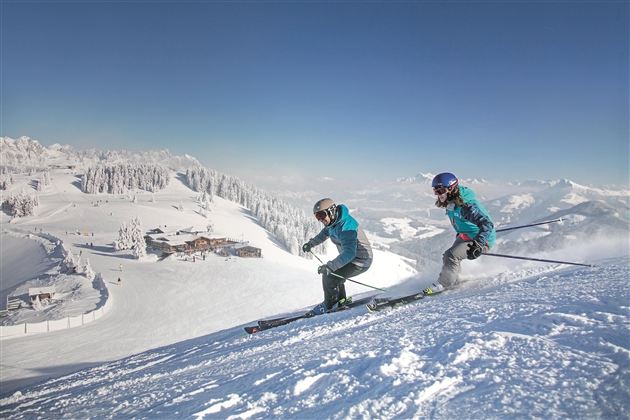 Zwei Skifahrer fahren einen verschneiten Hang hinunter. Im Hintergrund ist eine malerische Winterlandschaft zu sehen.