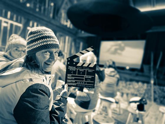 Eine fröhliche Person hält eine Klappe in einem Kino oder Filmraum. Im Hintergrund sind weitere Menschen und eine große Leinwand zu sehen.