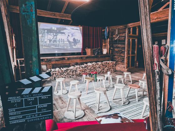 Ein gemütlicher Veranstaltungsraum mit Stühlen und einem großen Bildschirm. Der Raum hat eine rustikale Atmosphäre mit Holzelementen und einer Filmklappe im Vordergrund.