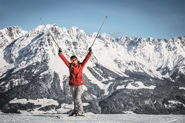 Eine glückliche Skifahrerin steht auf einem schneebedeckten Berg mit den Händen in die Luft. Im Hintergrund sieht man majestätische Berge und eine klare Winterlandschaft.