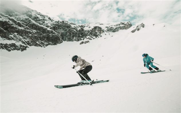 Zwei Skifahrer fahren über einen schneebedeckten Hang. Im Hintergrund sind beeindruckende Berge und ein bewölkter Himmel zu sehen.