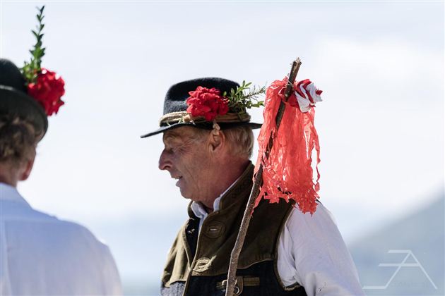 Een man in traditionele kleding met een bloeiende hoofdtooien en een stok. Op de achtergrond zijn vage silhouetten van andere personen te zien.