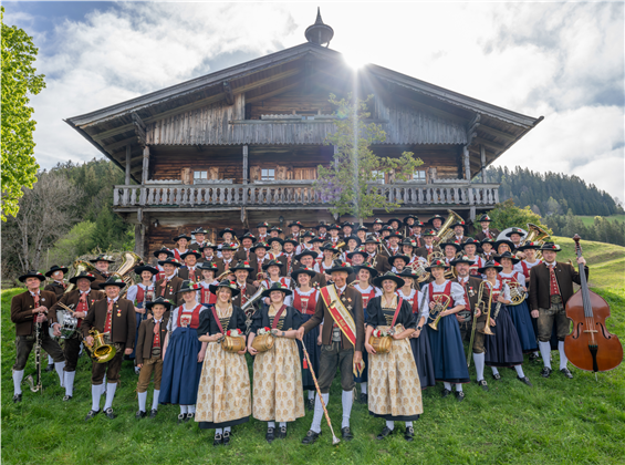 Eine große Gruppe von Menschen in traditioneller Tracht versammelt sich vor einem rustikalen, alpinen Holzhaus. Sie halten verschiedene Instrumente und stehen auf einer grünen Wiese.