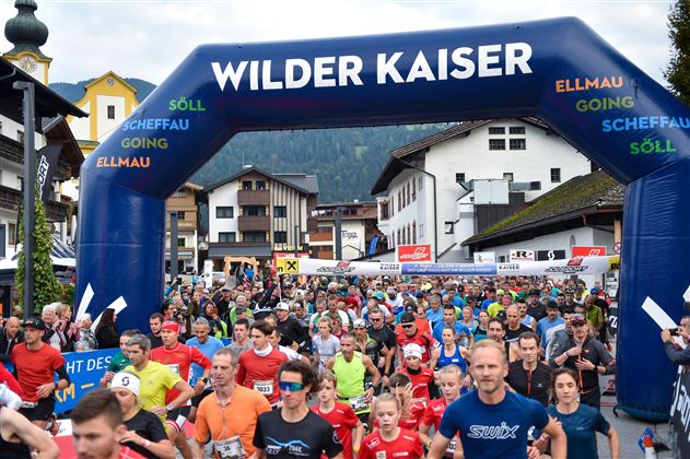 Ein großes Teilnehmerfeld beim Start eines Rennens unter einem blauen Tor mit der Aufschrift "Wilder Kaiser". Viele Läufer in bunten Shirts bewegen sich in Richtung des Starts in einer malerischen Umgebung.