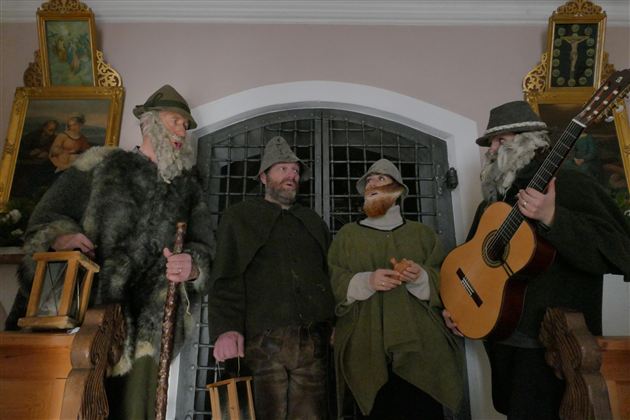 Eine Gruppe von vier Männern in historischen Kostümen steht vor einem Gittertor. Einer der Männer hält eine Gitarre und die anderen haben verschiedene Accessoires in der Hand.