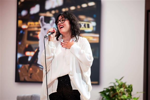 Eine Frau mit Brille singt lächelnd ins Mikrofon. Im Hintergrund sind unscharfe Lichter und eine Pflanze zu sehen.