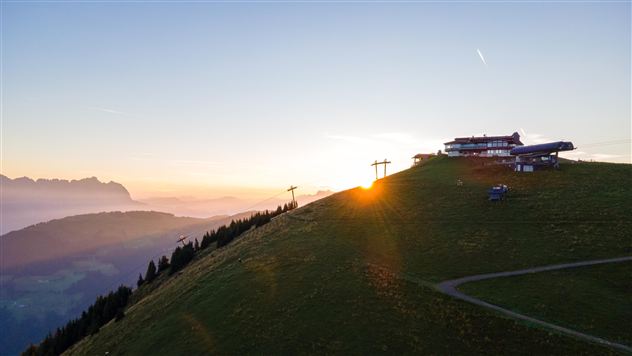 Eine malerische Berglandschaft bei Sonnenaufgang mit einem Gebäude und Seilbahnen. Sanfte Hügel und Wälder umgeben die Szenerie.