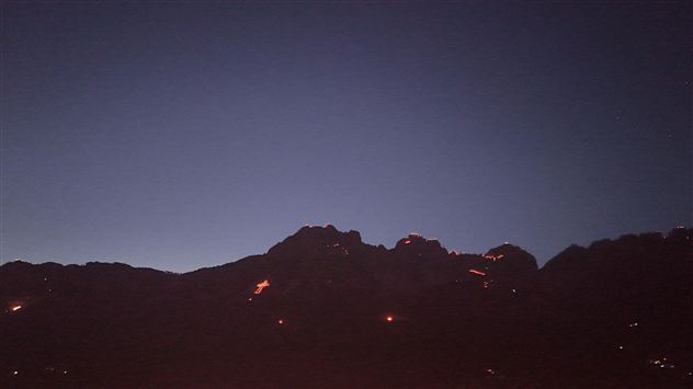 Ein gemütliches Berghaus aus Holz mit einer großen Terrasse. Umgeben von grünen Wiesen und Bäumen unter klarem Himmel.