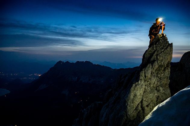 A climber stands on a high rock and inspects his gear. In the background, the mountainous landscapes and a clear sky at dusk are visible.