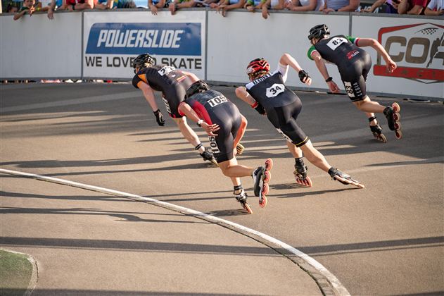 Vier Rollschuhfahrer sprinten in schneller Formation auf einer Rennbahn. Im Hintergrund ist eine Menschenmenge zu sehen, die das Rennen anfeuert.