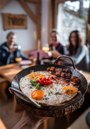 Srdcové snídaně se smaženými vejci, slaninou a čerstvými bylinkami v rustikální pánvi. V pozadí jsou veselí hosté, kteří si vychutnávají nápoje.