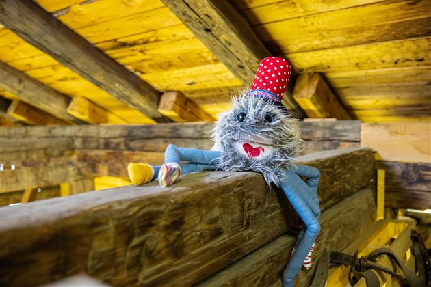 Ein lustiges Stofftier mit einem roten Hut sitzt auf einem Balken. Es hat einen fröhlichen Gesichtsausdruck und farbenfrohe Füße.