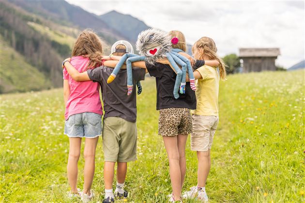 Vier Kinder stehen auf einer Wiese und umarmen sich. Eines der Kinder hält ein kuscheliges Spielzeug in Form eines Monsters.