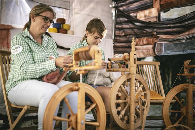Zwei Frauen sitzen an Spinnrädern und arbeiten mit Wolle. Sie tragen kariert gestreifte Blusen und konzentrieren sich auf ihre Handarbeit.