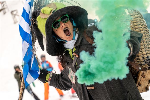 Een persoon draagt een grote hoed en zonnebril en steekt een groene rookfakkel af. Op de achtergrond zijn mensen op ski's te zien.