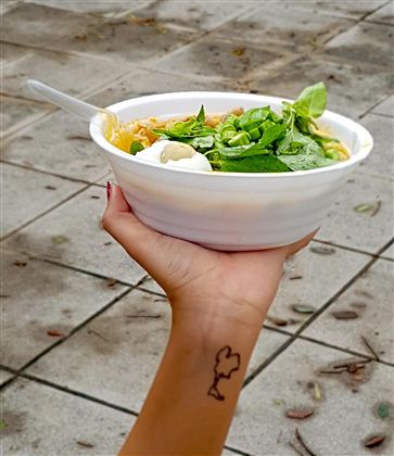 Eine Hand hält eine Schüssel mit frischen Nudeln und grünem Blattgemüse. Im Hintergrund sind Pflastersteine zu sehen.