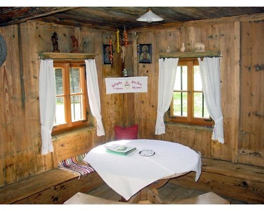 A cozy room with wooden walls and two windows. A round table with a white tablecloth stands in the middle, surrounded by bench cushions.