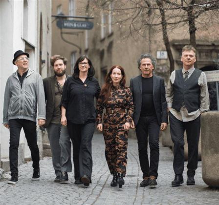 Eine Gruppe von sechs Menschen steht auf einer steinernen Straße. Sie tragen verschiedene Outfits und wirken entspannt.