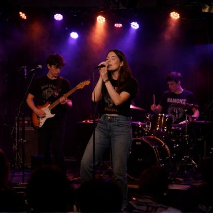 Eine Band spielt auf einer Bühnen mit bunten Lichtern. Die Sängerin steht im Mittelpunkt, während die anderen Musiker aktiv mitspielen.