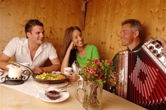 Eine fröhliche Runde von drei Personen in einem rustikalen Raum. Der Mann spielt Akkordeon, während das Paar mit Essen und Getränken genießt.