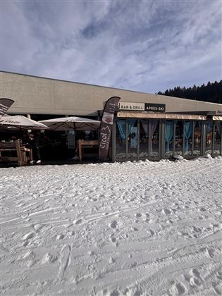 An après-ski bar and grill with a terrace, surrounded by snow. In the background, there are trees and a clear sky.