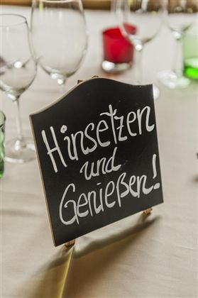 Ein Schild mit der Aufschrift „Hinsetzen und Genießen!“ steht auf einem Tisch. Im Hintergrund sind Gläser und Kerzen sichtbar.