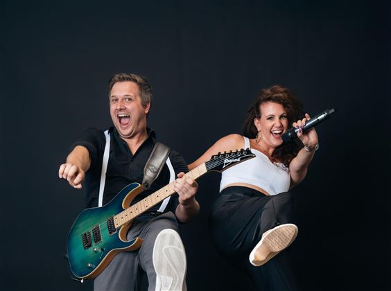 Ein Musiker mit einer Gitarre und eine Sängerin posieren fröhlich vor einem neutralen Hintergrund. Beide strahlen Energie und Freude aus.